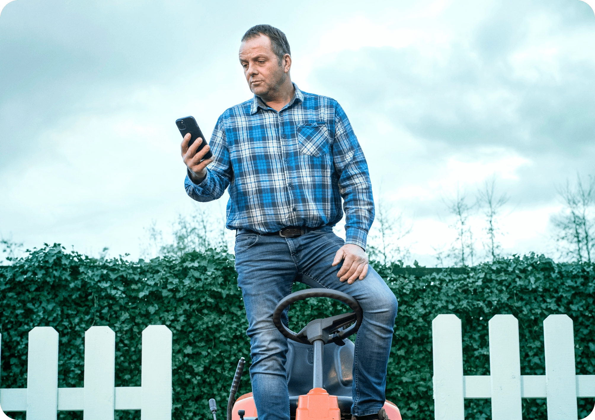 Mann steht auf Rasentraktor im Freien, schaut auf Smartphone unter bewölktem Himmel.